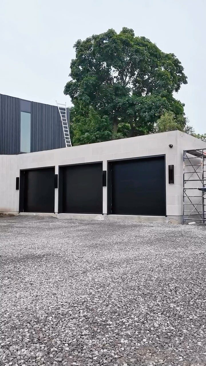 3 Car Garage Design: 8 Ideas for Style and Function 8 A modern three-car garage with black doors and a gravel driveway under a large green tree.