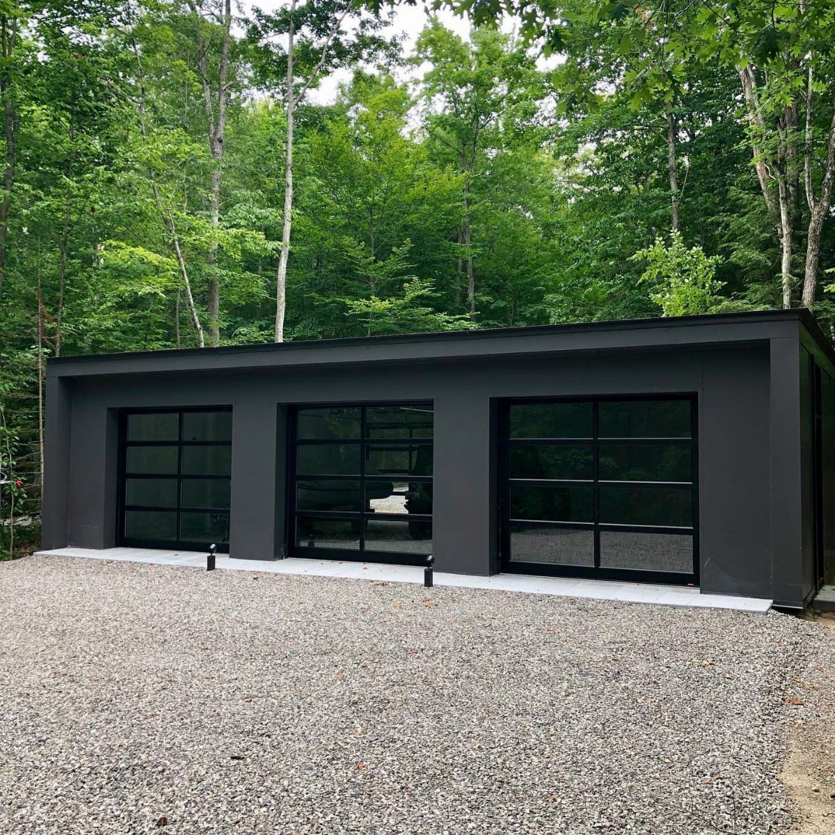 3 Car Garage Design: 8 Ideas for Style and Function 3 A modern dark grey three-car garage with black glass doors set against a dense green forest.