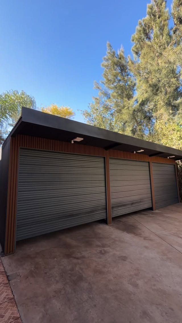 3 Car Garage Design: 8 Ideas for Style and Function 5 A modern three-car garage with dark grey roller doors and wood paneling accents under a blue sky.