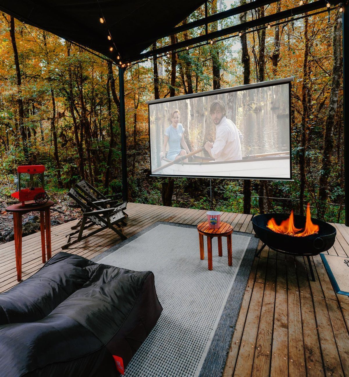 Outdoor movie screen playing a film on a wooden deck surrounded by autumn trees.