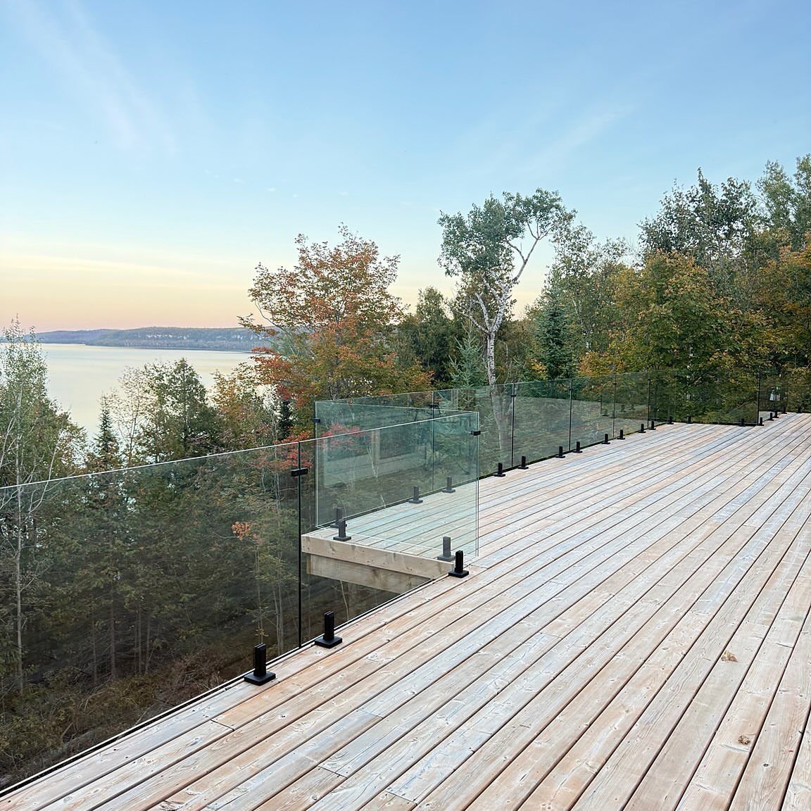 A wooden deck with clear glass railings overlooking a lake and lush green forest.