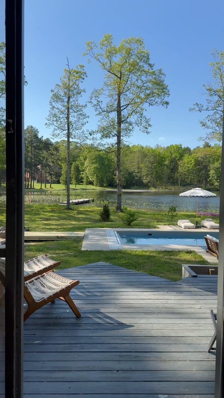 Wooden deck with lounge chairs overlooking a swimming pool and a calm lake surrounded by trees.