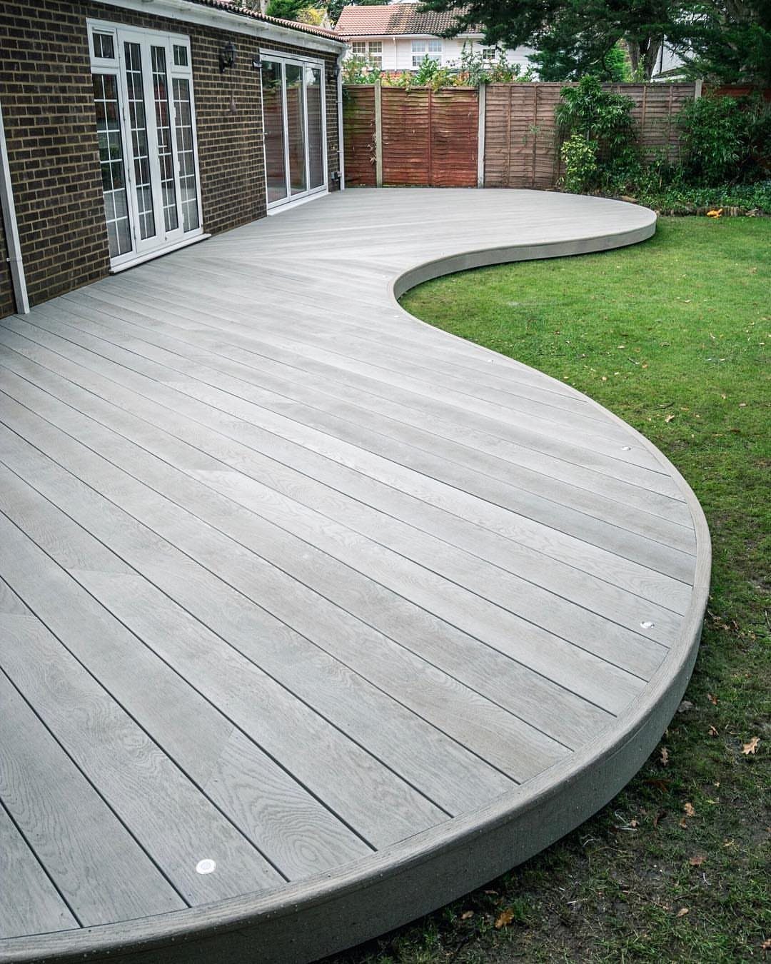 A large, light grey curved wooden deck extending from a brick house onto a green lawn.