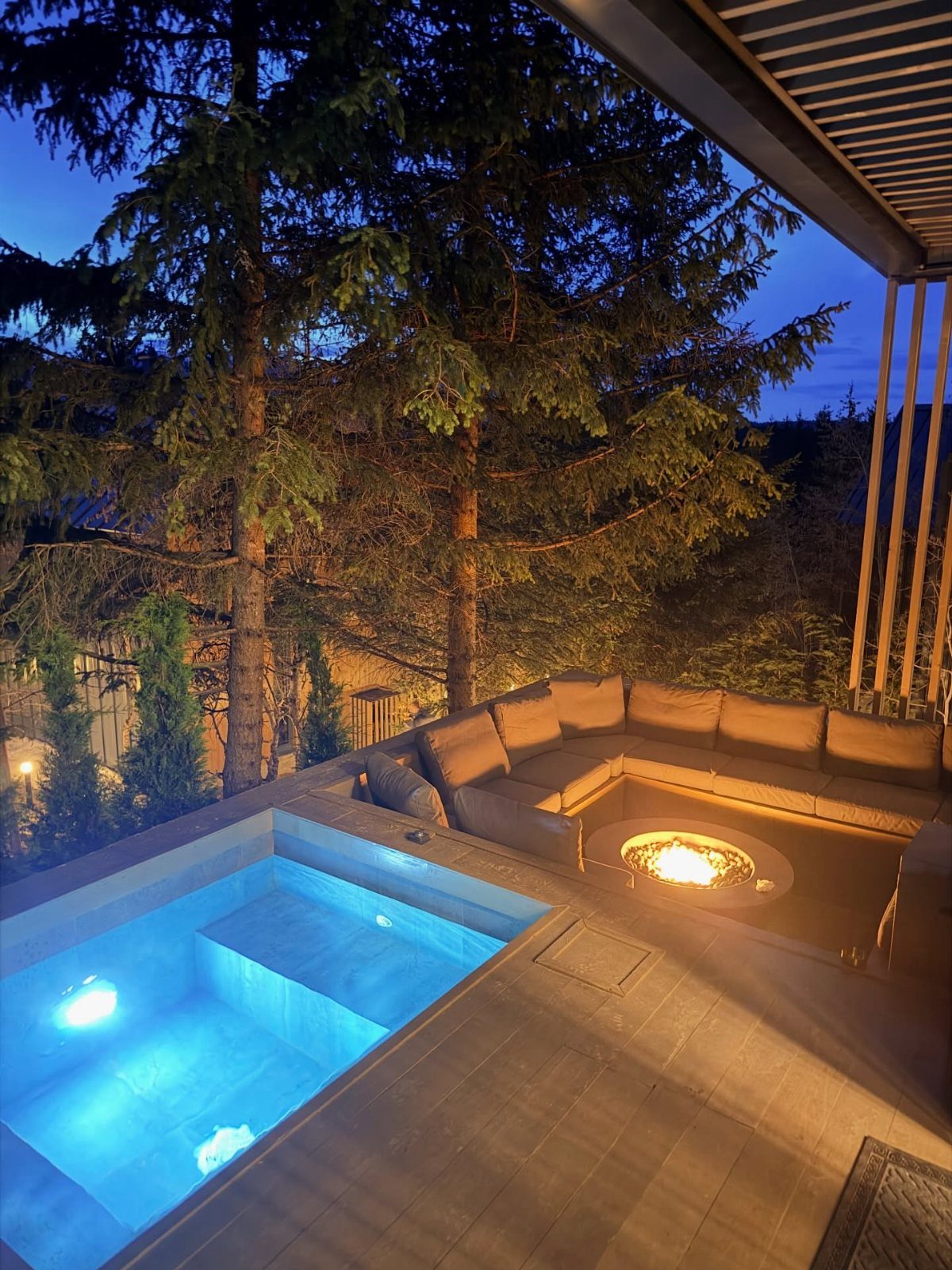 A luxury deck at dusk with a glowing blue hot tub and a warm fire pit.