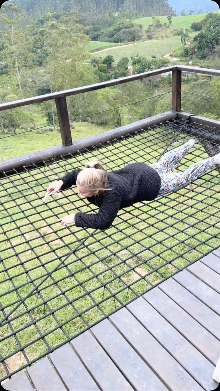 A person lying on a large black rope net installed on a wooden deck overlooking a green valley.