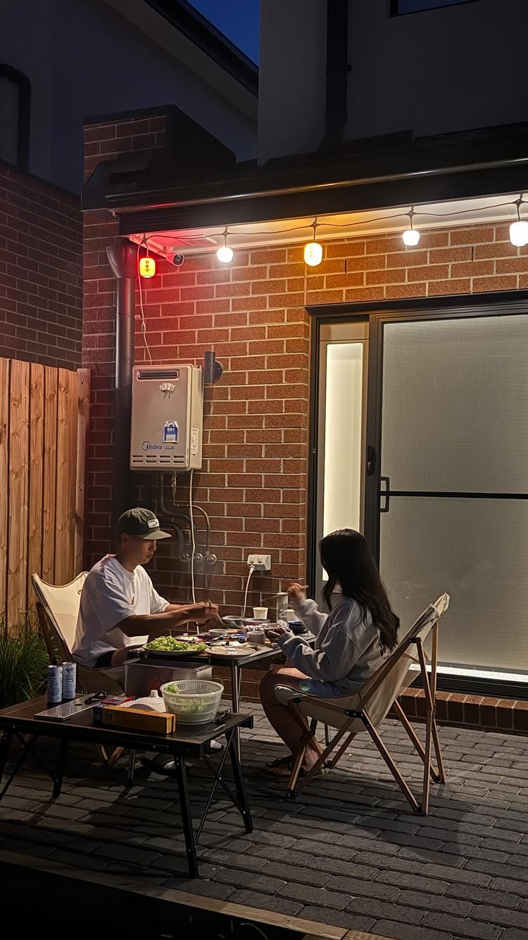 12 Backyard BBQ Ideas for Grill Area and Patio Layout 7 Two people dining at a small table on a brick patio under glowing string lights.