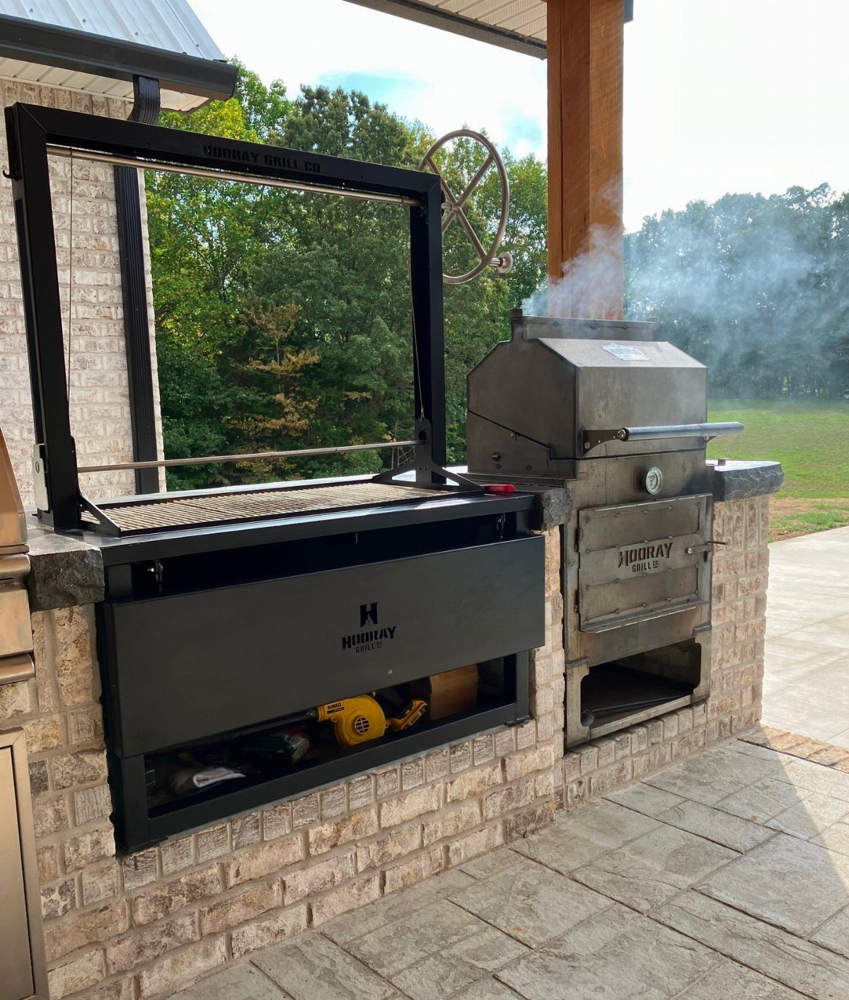 A stainless steel built-in smoker emitting smoke on a brick outdoor kitchen counter.