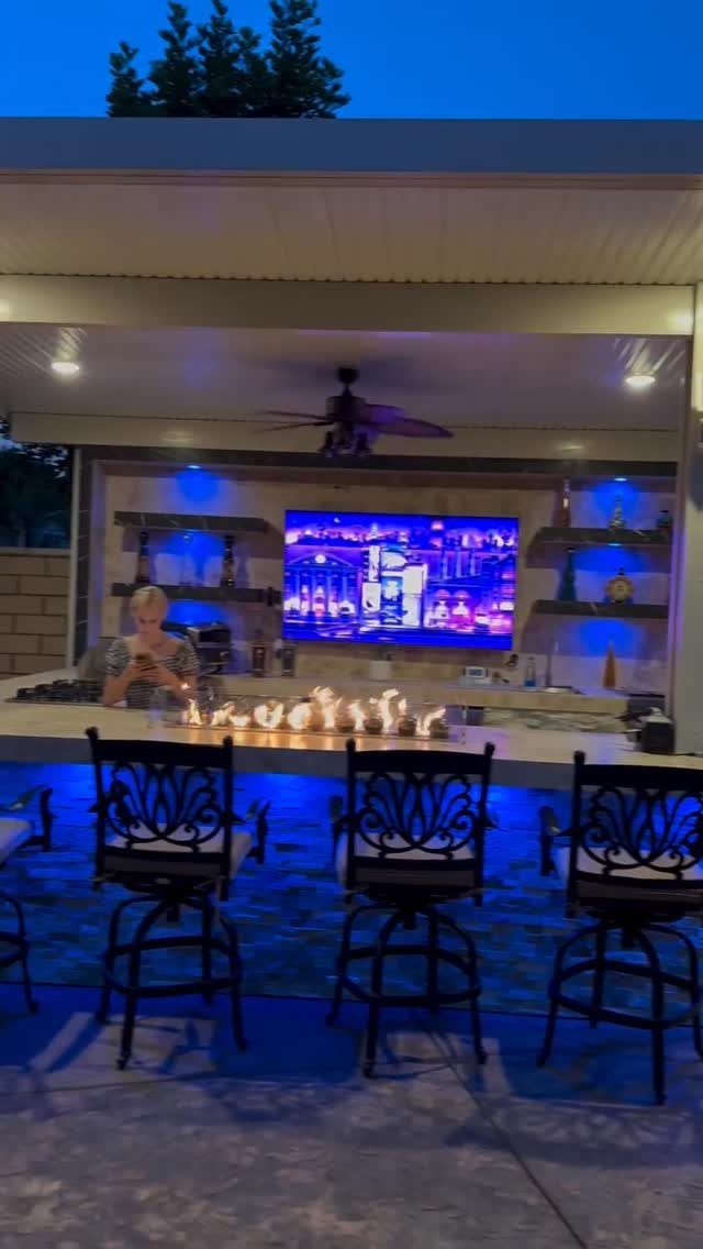 A linear fire pit built into an outdoor kitchen counter with blue lighting and bar stools.