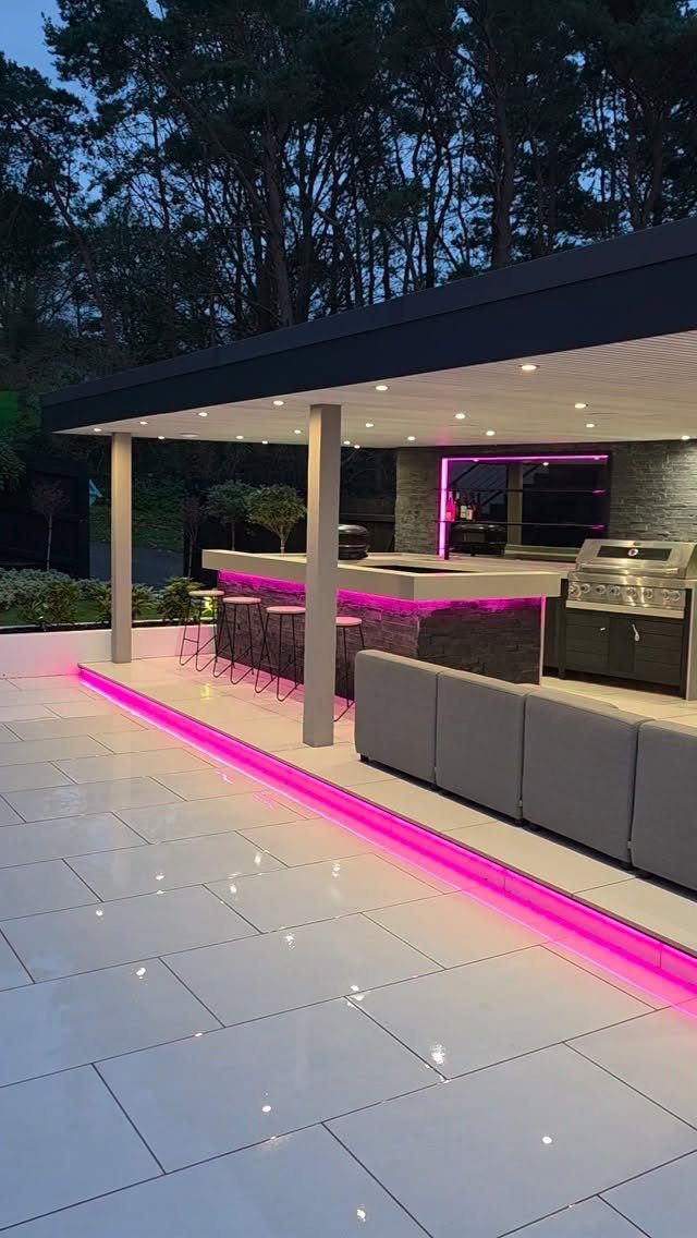 Modern outdoor kitchen with pink LED lighting, stone counters, and a built-in grill under a pergola.
