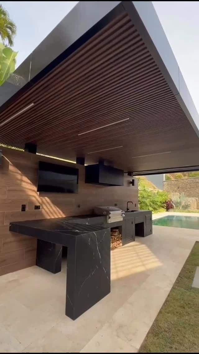 Modern outdoor kitchen with black marble counters, built-in grill, and TV next to a swimming pool.