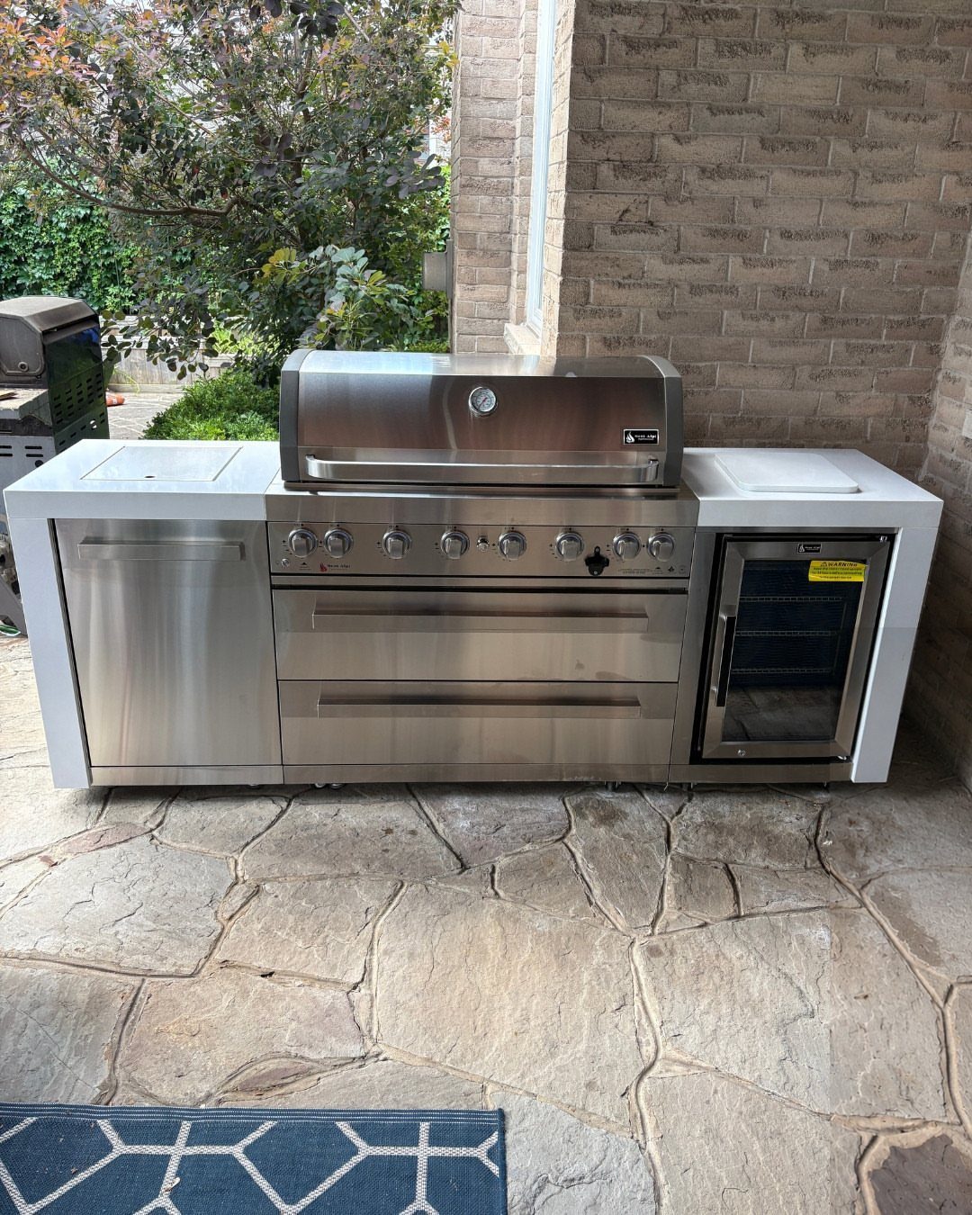 Stainless steel outdoor grill station with built-in drawers and a small refrigerator on a stone patio.