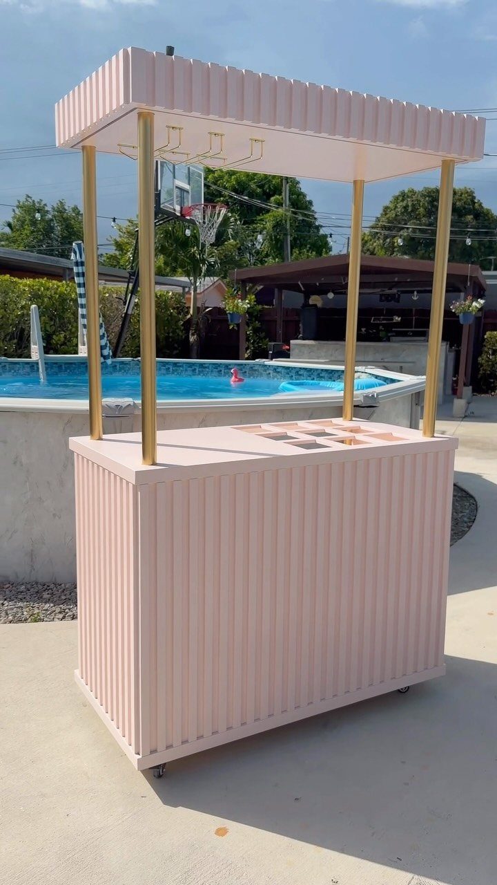 Backyard Pool Bar Ideas: 10 Upgrades for Summer Entertaining 3 A pink fluted portable bar with gold poles stands on a patio near a pool.