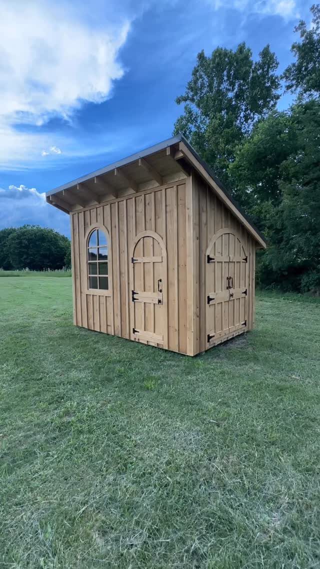 Board and Batten Shed: 5 Color Ideas That Work 3 A wooden garden shed with vertical siding and arched doors sitting on a grassy field.