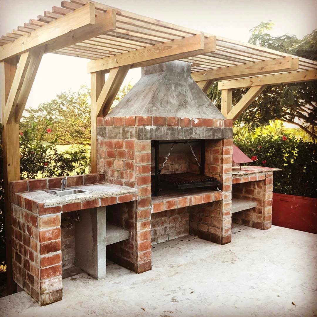 10 Brick BBQ Ideas: From Classic Pits to Pizza-Oven Combos 3 A large brick outdoor barbecue station with a chimney and sink under a wooden pergola.