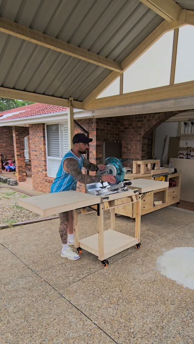 A man using a miter saw on a wooden mobile workbench with wheels under a carport.