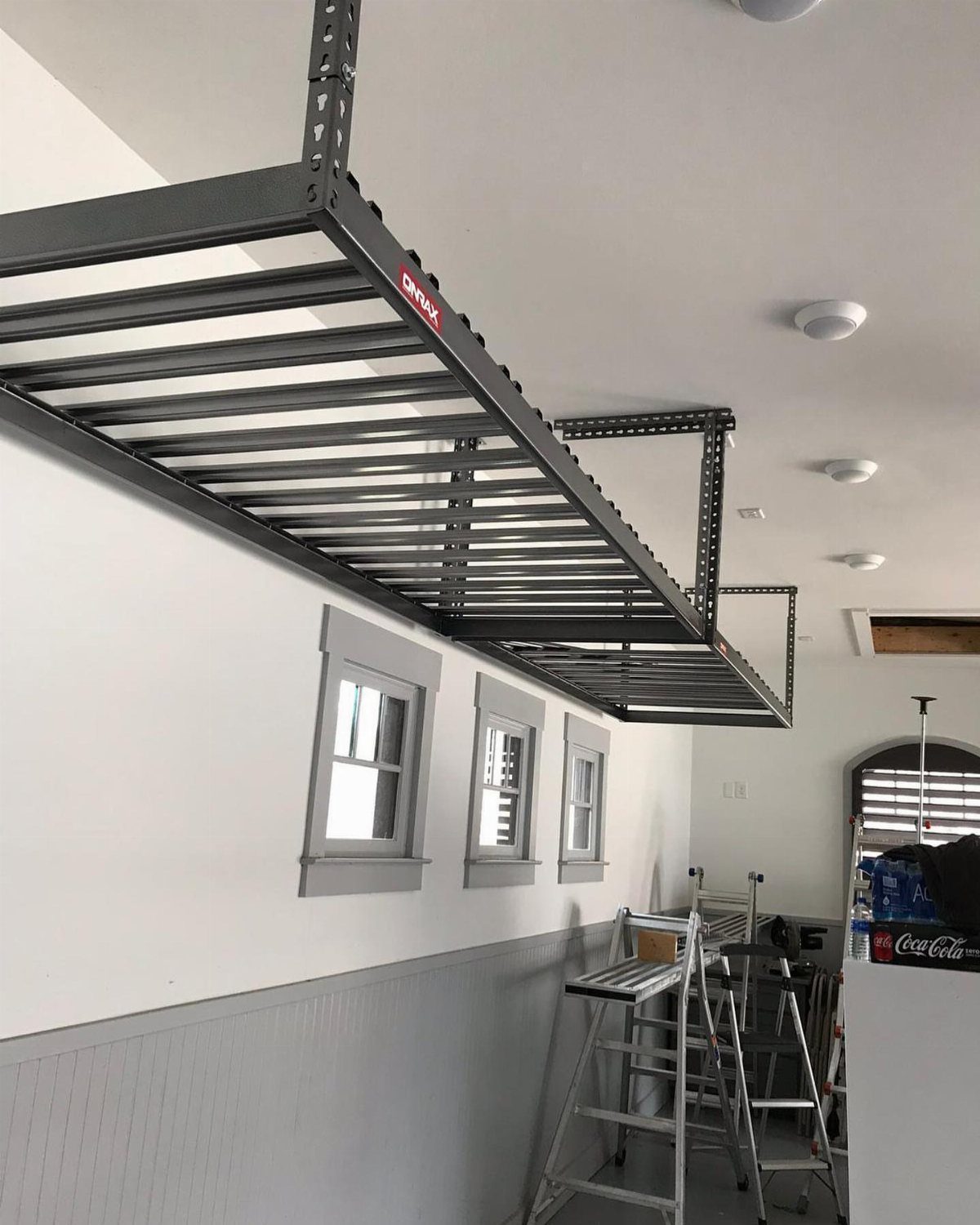 Two grey metal overhead storage racks mounted to a white garage ceiling above windows.