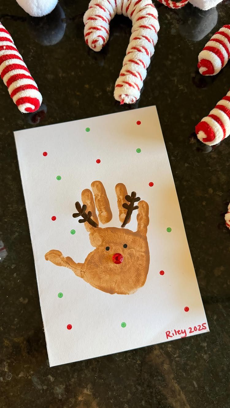A brown handprint reindeer painting with red nose and antlers on white paper surrounded by candy canes.