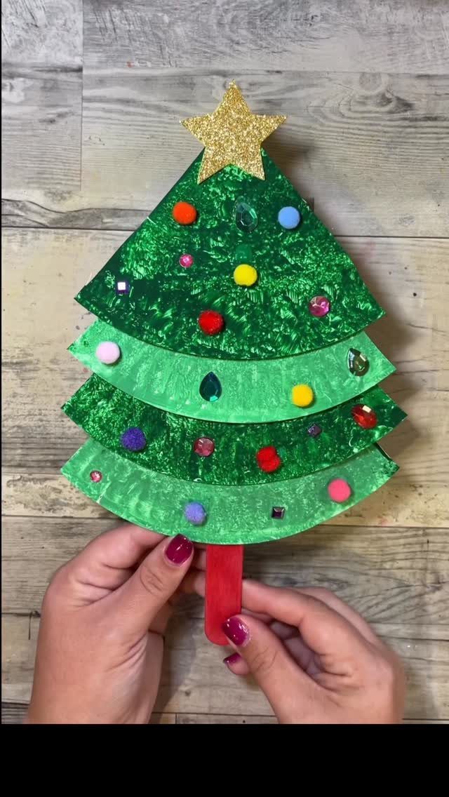 Hands holding a handmade green paper Christmas tree decorated with colorful pom-poms and a gold star.