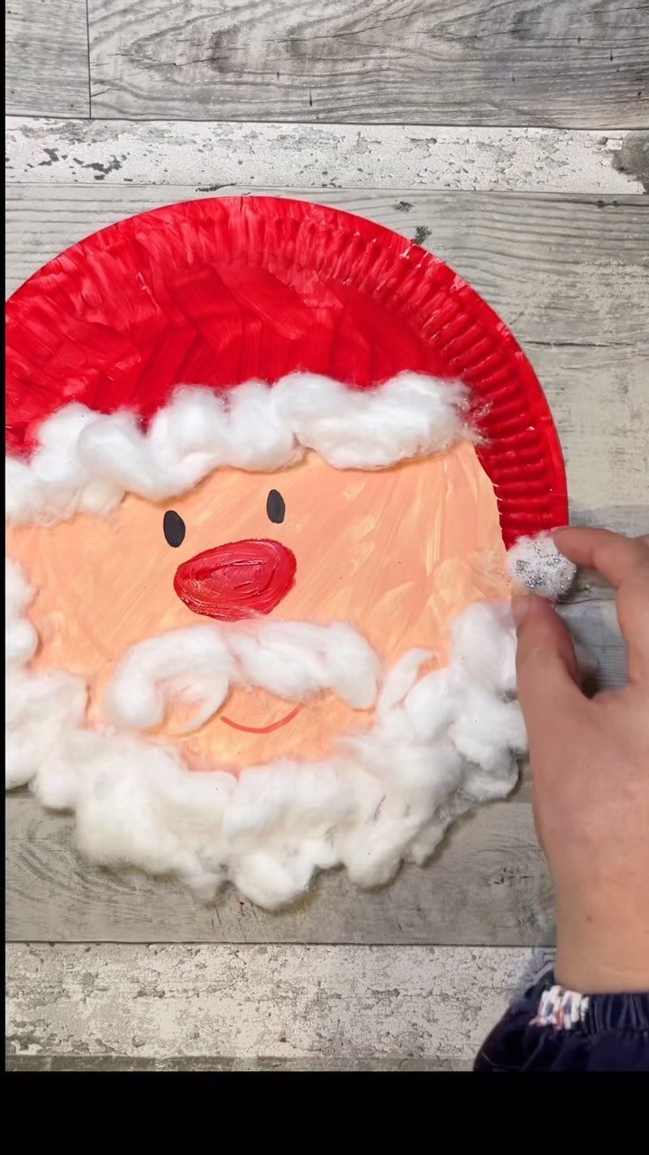 A hand placing white cotton balls onto a paper plate Santa Claus craft.