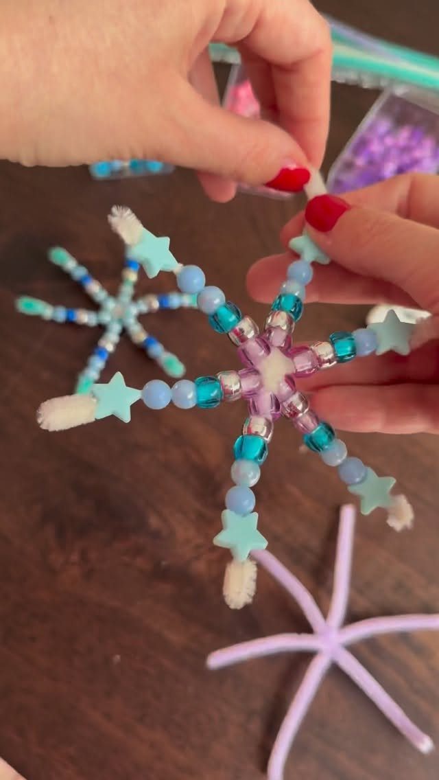 Hands assembling a snowflake ornament made from blue beads, white pipe cleaners, and small stars.