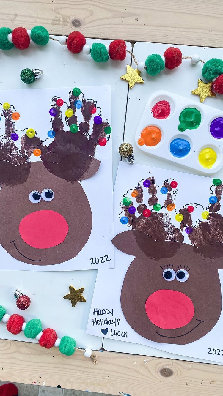 12 Christmas Family Craft Ideas: Projects for All Ages 9 Two handmade reindeer Christmas cards made with brown handprints and colorful pom-pom antlers on a wooden table.