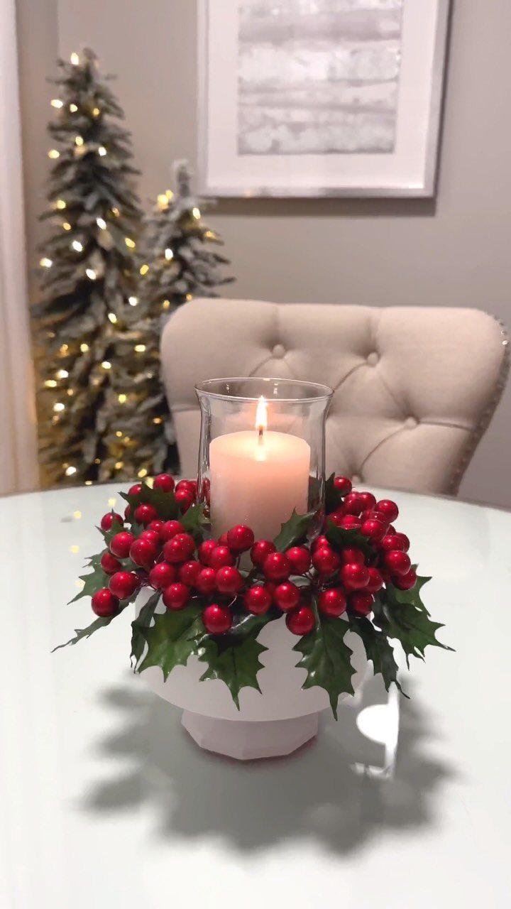 A lit white candle surrounded by red berries and holly leaves in a white bowl.