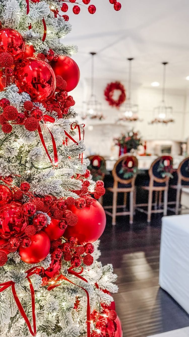 Flocked Christmas tree decorated with red ornaments, red berries, and red ribbons in a bright room.