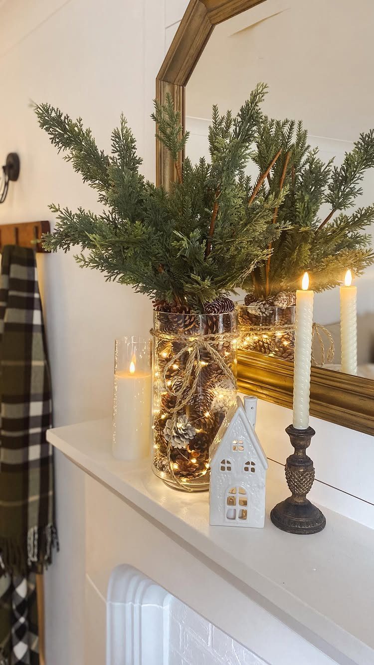 Evergreen branches in a glass vase with fairy lights on a white mantelpiece.