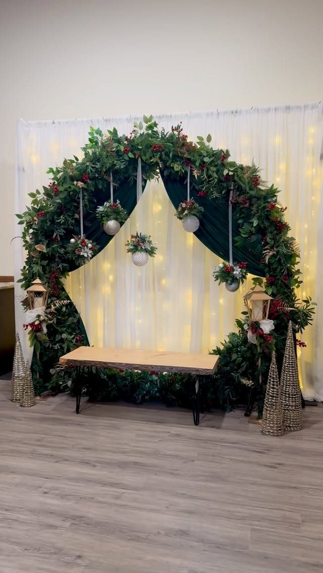 A lush green foliage arch decorated with red berries, hanging ornaments, and lanterns over a wooden bench.