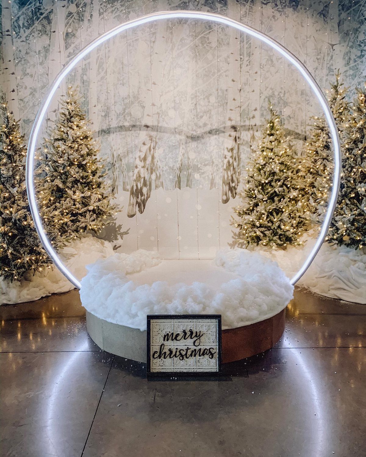 A circular lighted Christmas photo booth backdrop with snowy trees and a merry christmas sign.