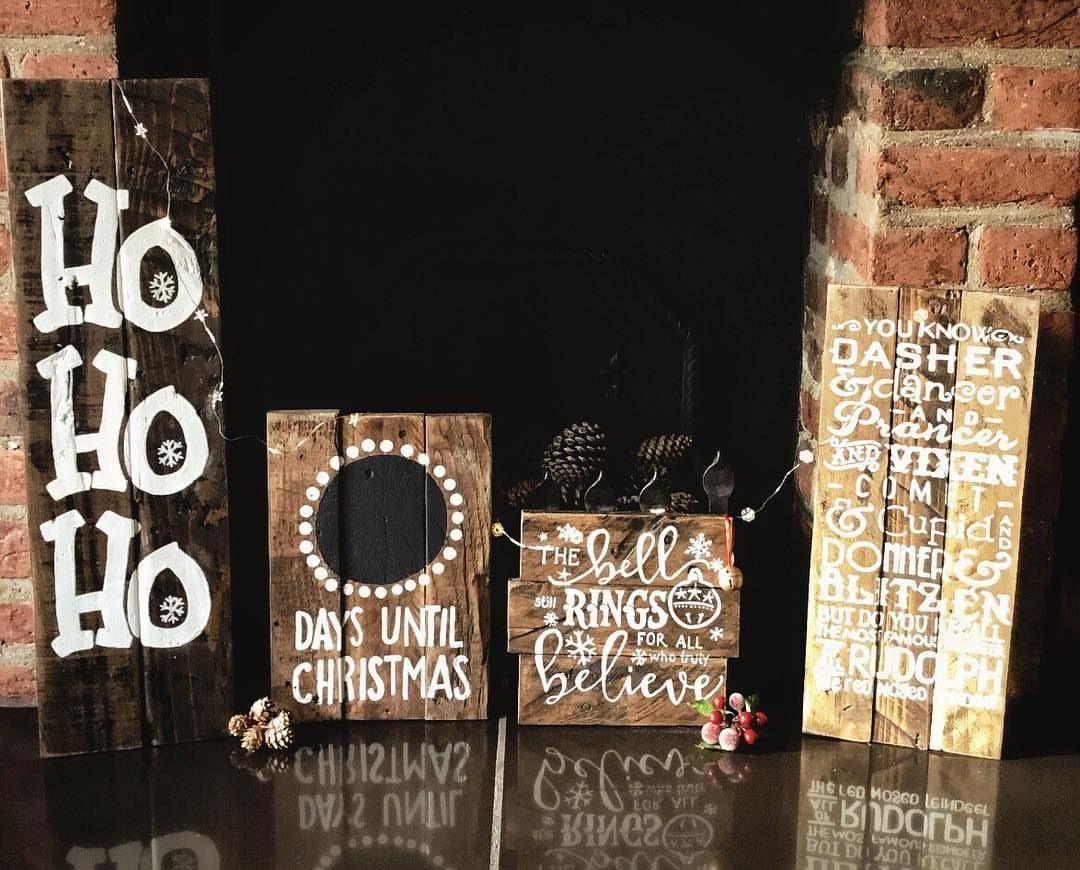 Four rustic wooden Christmas signs with white lettering standing in front of a dark fireplace.