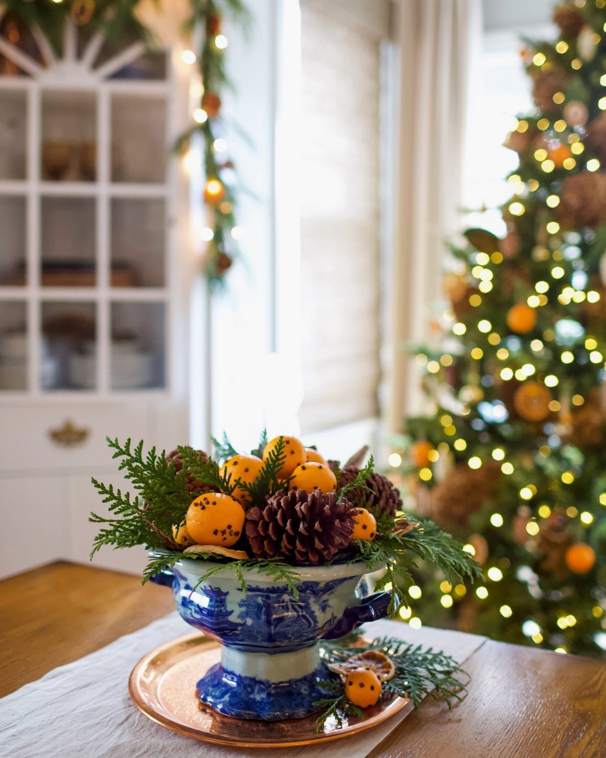 Orange pomanders and pinecones arranged with evergreen sprigs in a blue ceramic bowl.