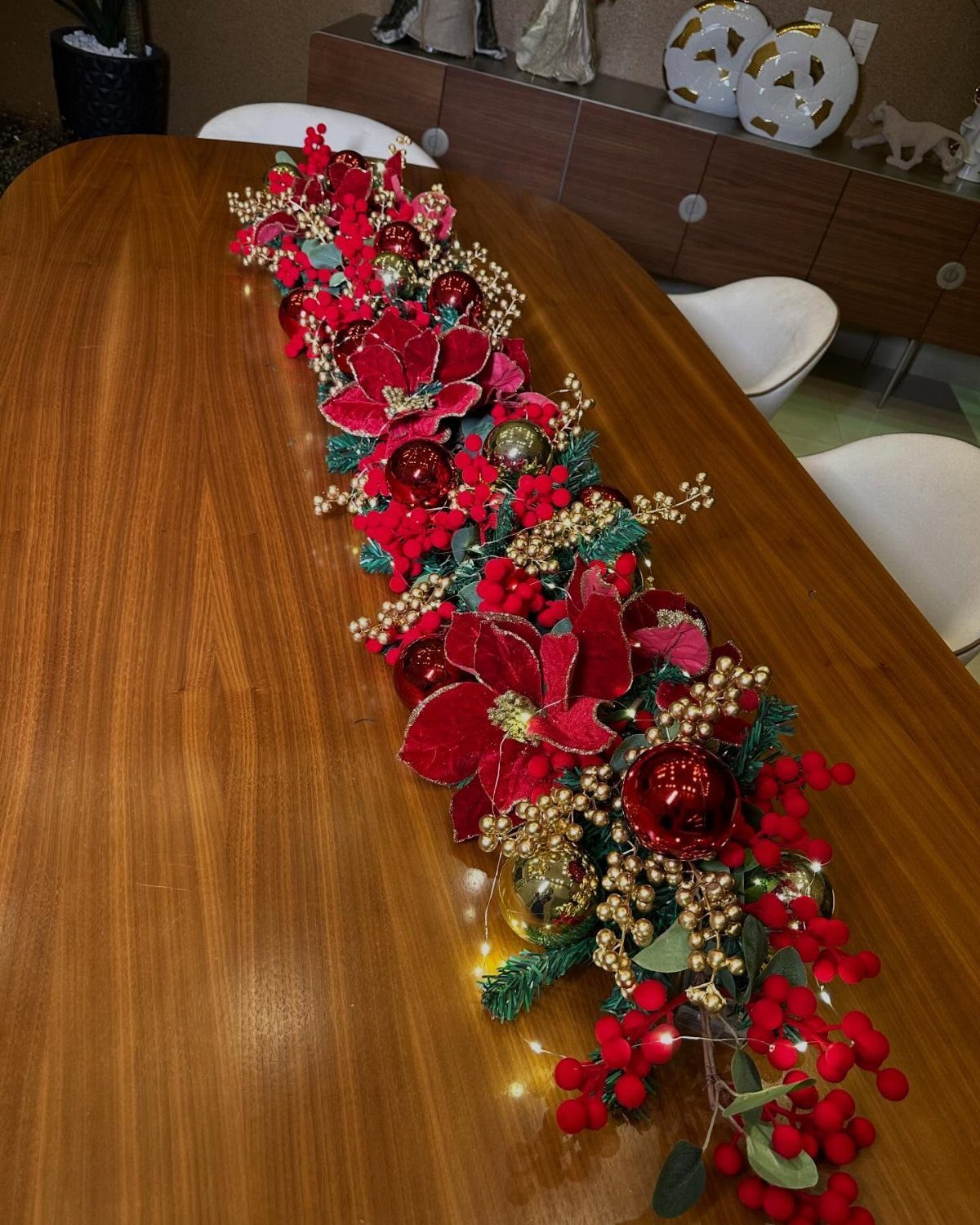 Red poinsettias, gold ornaments, and red berries forming a long Christmas table centerpiece runner.