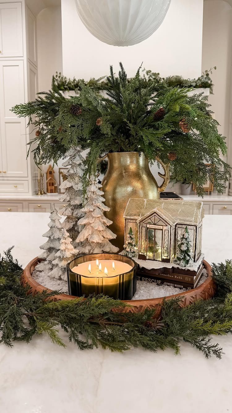 Christmas centerpiece with evergreen branches, gold pitcher, frosted trees, and a glowing candle on marble.