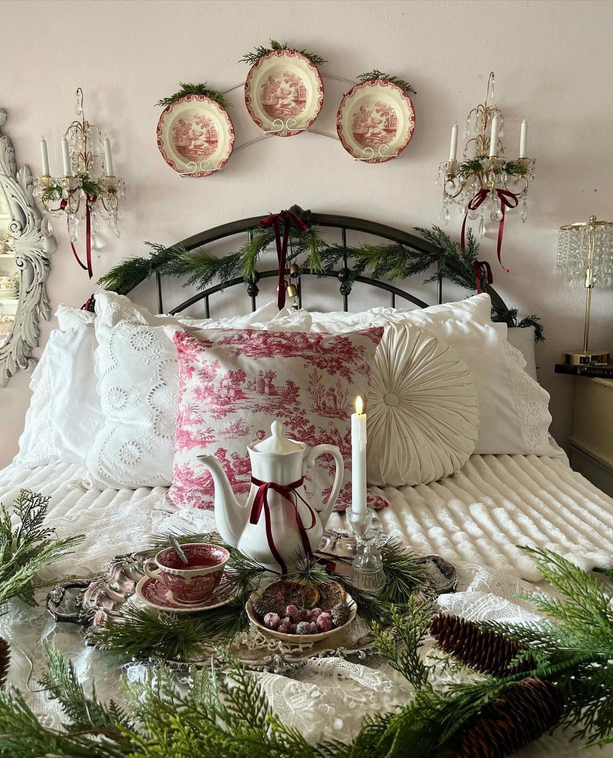 Decorated bed with pink patterned pillows, white teapot, and evergreen garlands on a tray.