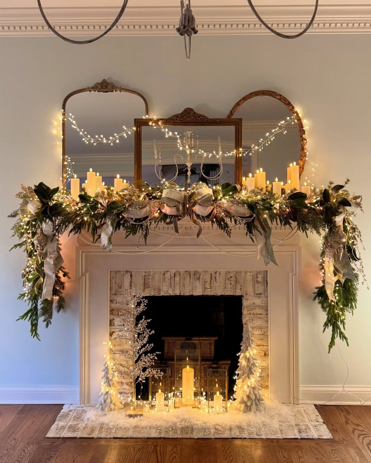 A decorated white fireplace mantel with green garland, white candles, and fairy lights under three mirrors.