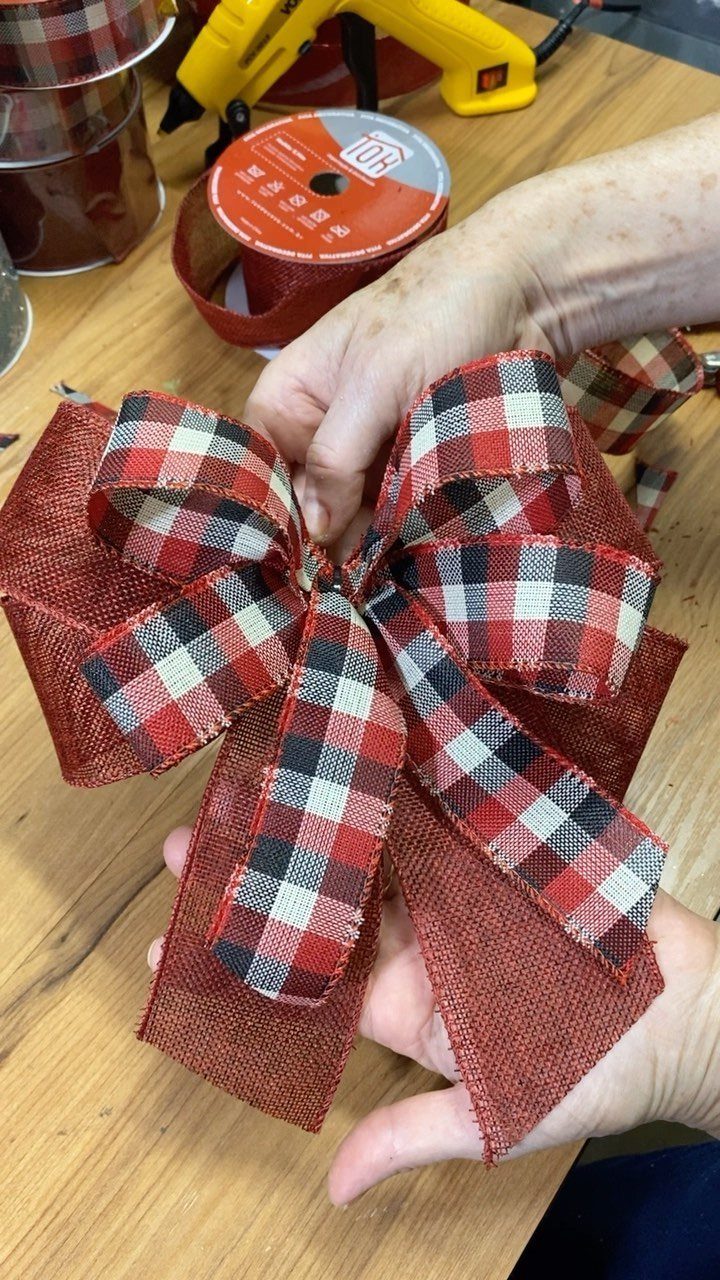 Hands holding a large decorative bow made of red burlap and plaid ribbon on a wooden table.