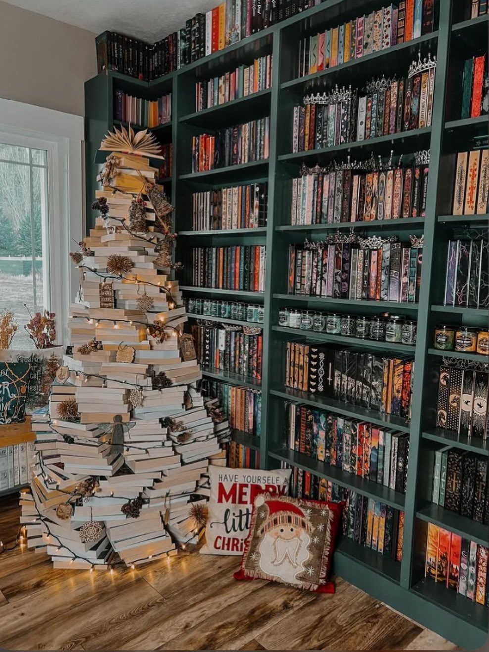 A Christmas tree made of stacked books decorated with lights and ornaments next to a bookshelf.