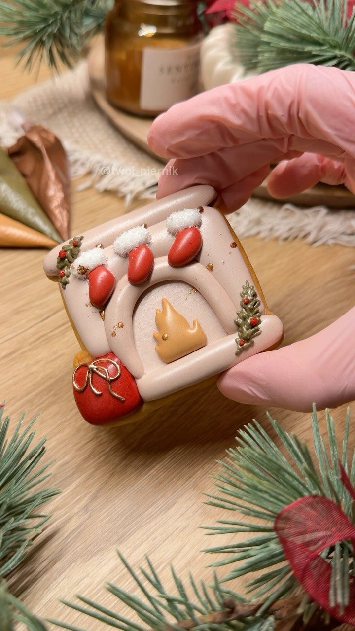 A person in pink gloves holding a decorated fireplace-shaped Christmas cookie with red stockings.