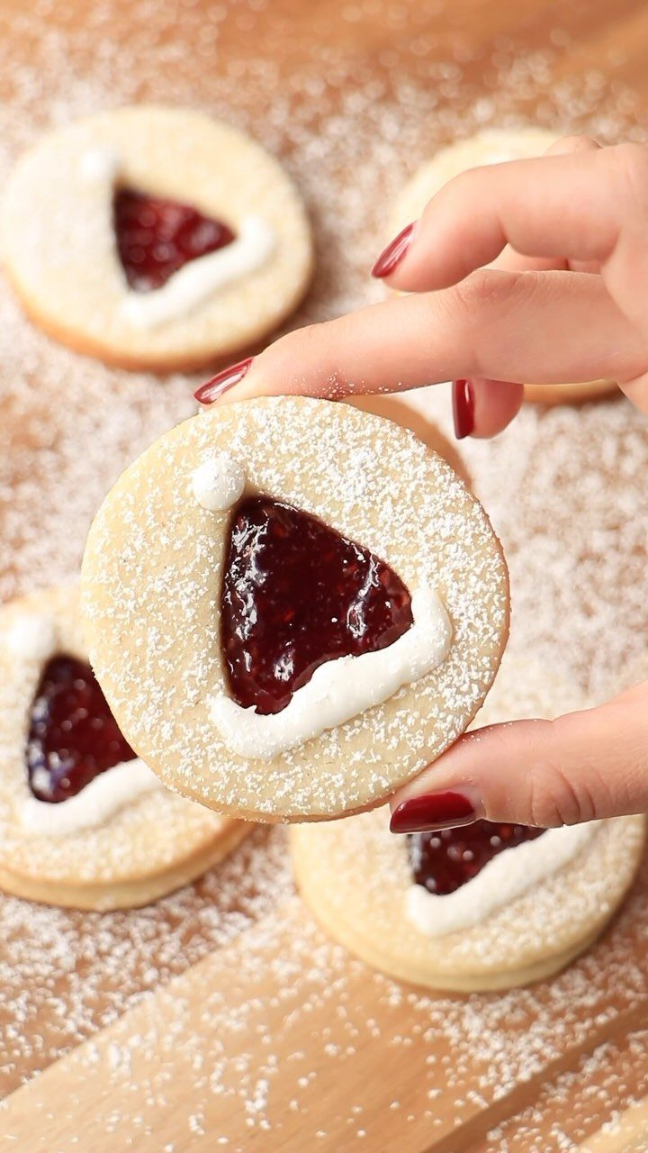 A hand holding a round jam-filled cookie with white icing on a powdered sugar surface.