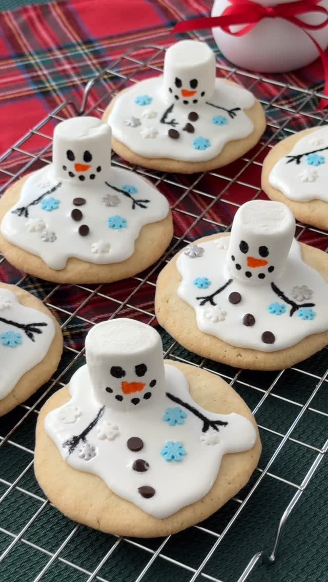 Decorated sugar cookies shaped like melting snowmen with marshmallow heads on a wire cooling rack.