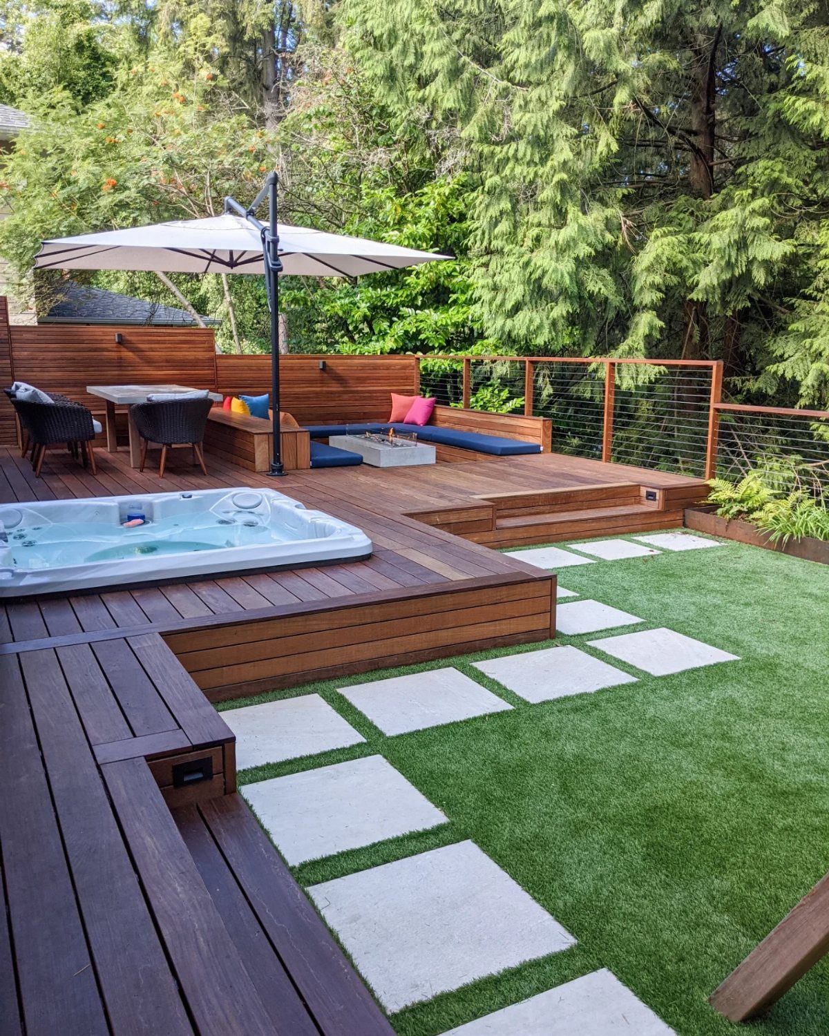 A multi-level wooden outdoor deck with a hot tub, dining area, and stepping stones on grass.
