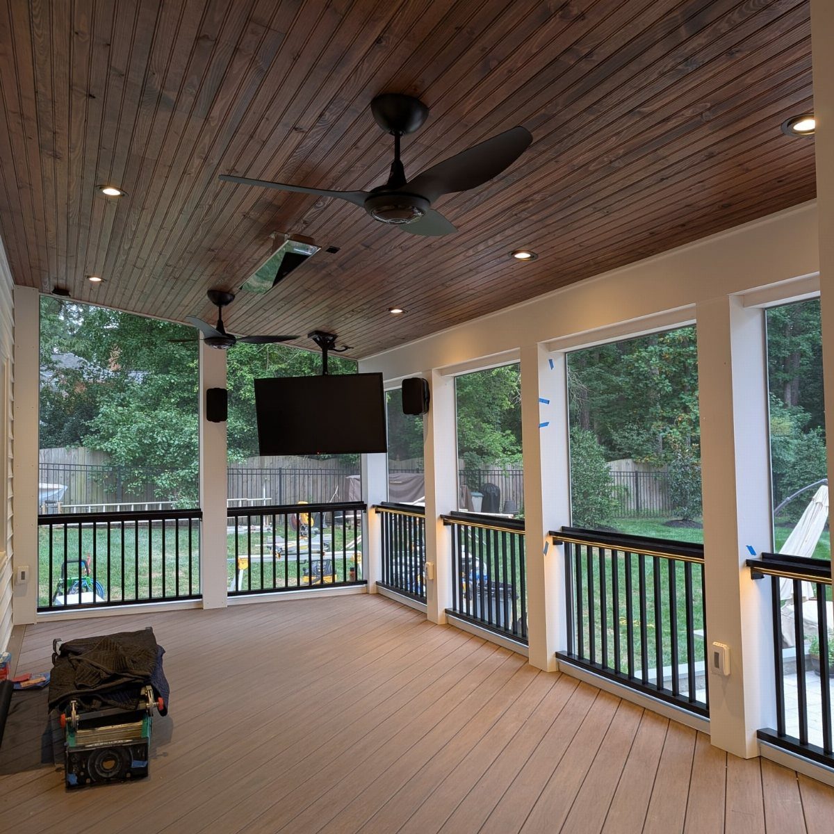 A covered outdoor wooden deck featuring ceiling fans, a television, and black railings overlooking a yard.