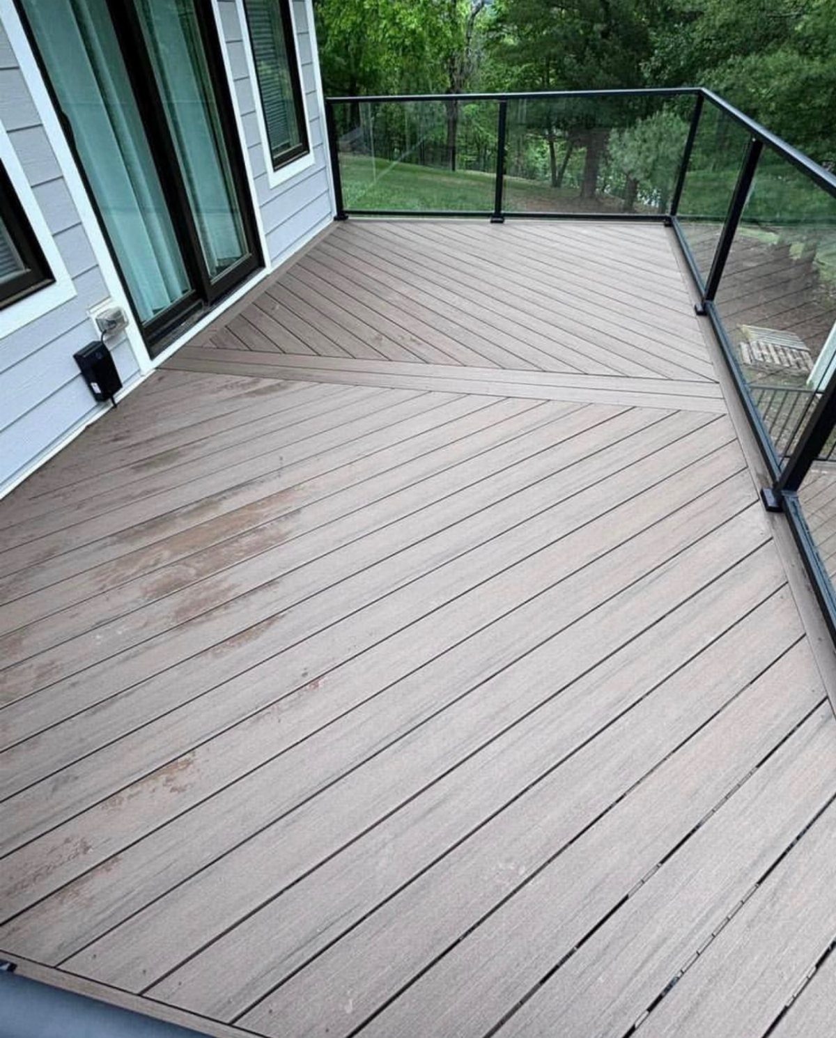 A wooden deck featuring a diagonal plank layout design with glass railings and green trees.