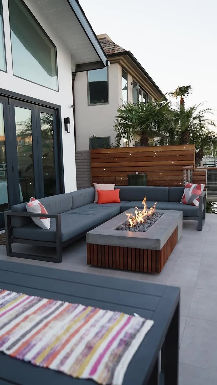 A modern grey sectional sofa surrounds a rectangular concrete fire pit with active flames on a patio.