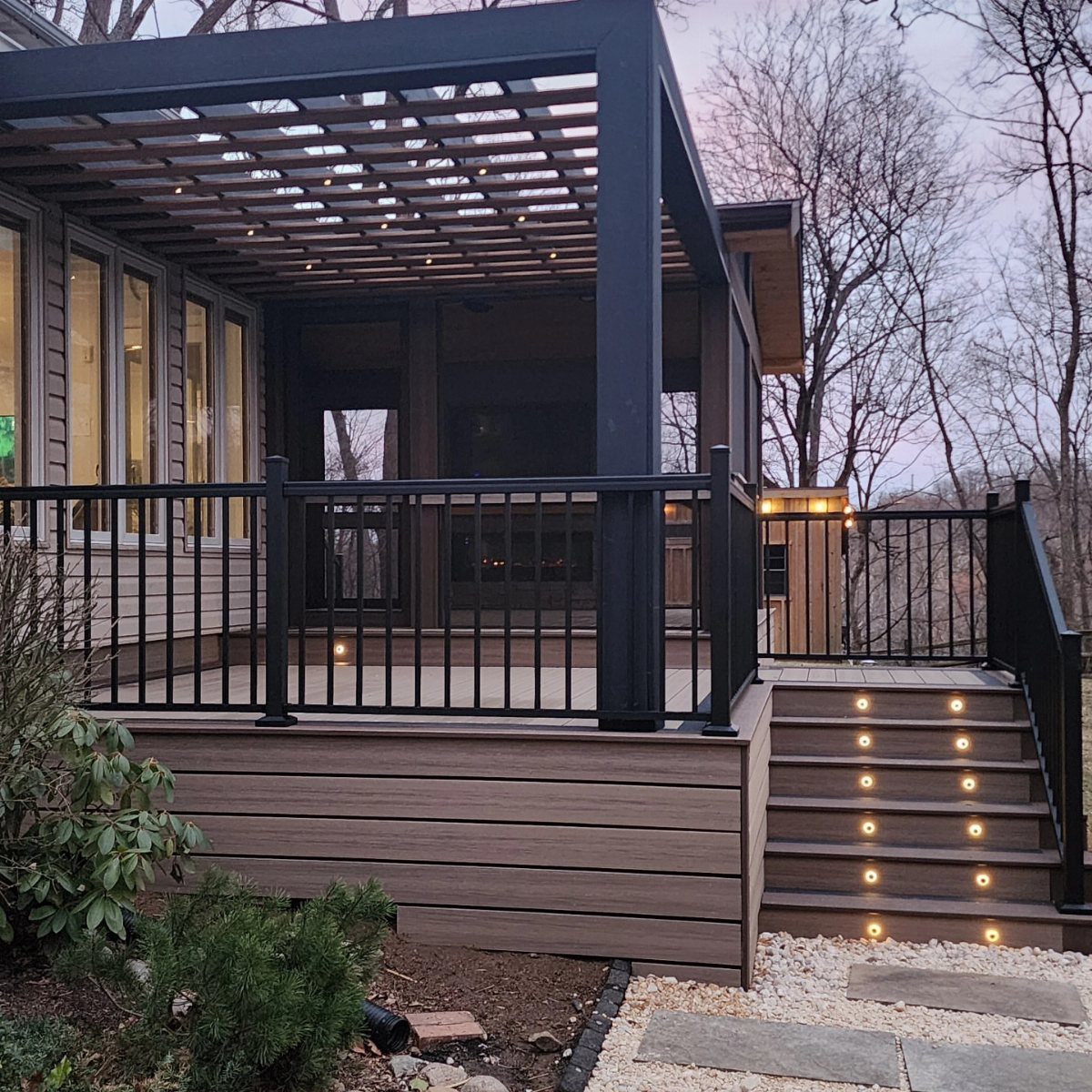 A modern wooden deck with a black pergola, railing, and illuminated stairs overlooking trees.