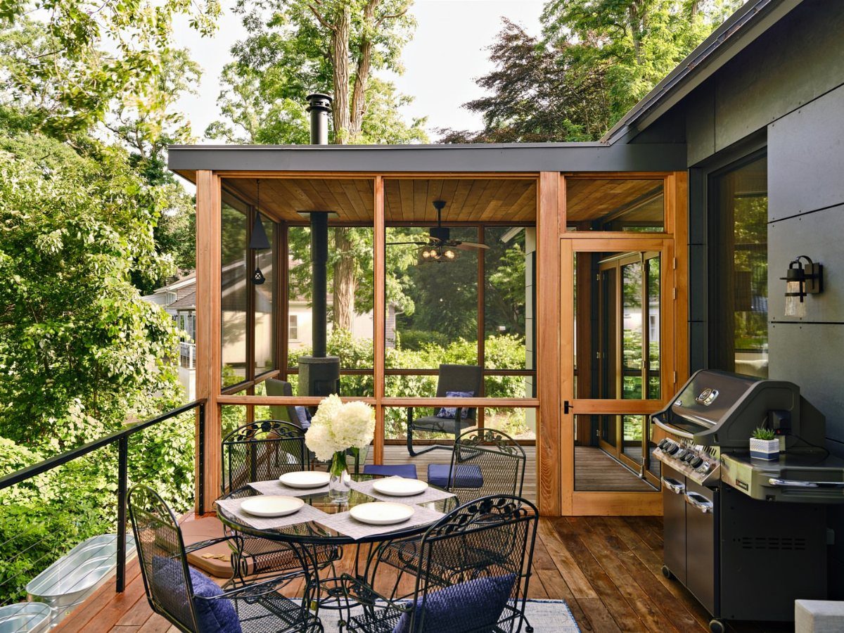 A wooden screened-in porch with a ceiling fan overlooking a lush green forest and outdoor deck.