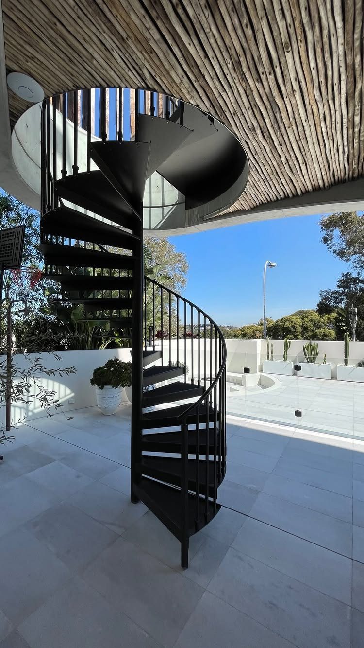 A black metal spiral staircase on a white tiled patio under a wooden pergola.