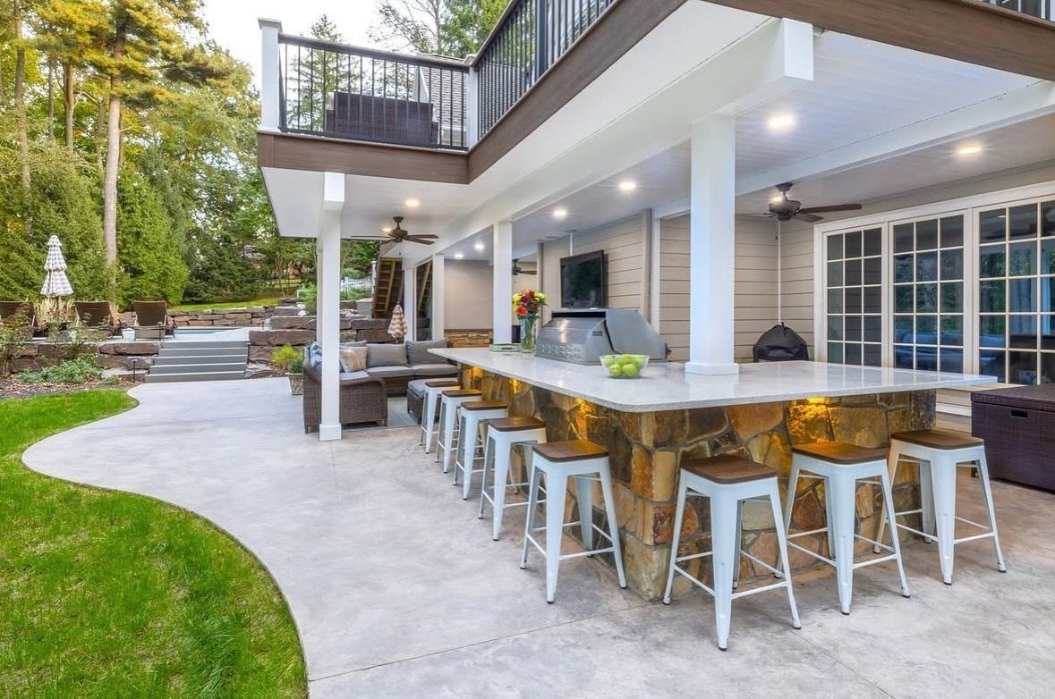 An outdoor kitchen with a stone bar, white stools, and a grill under a deck.