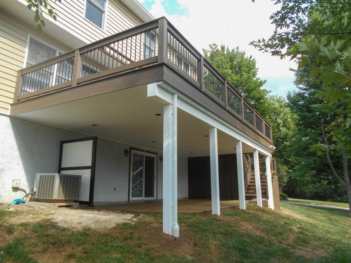 A large brown wooden deck with white support posts covering a concrete patio area below a house.